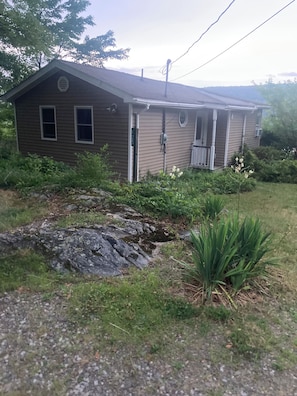 Une maison de plain-pied avec un porche à l’avant, un petit jardin agrémenté de rochers et de plantes, et un coffret de raccordement apparent.