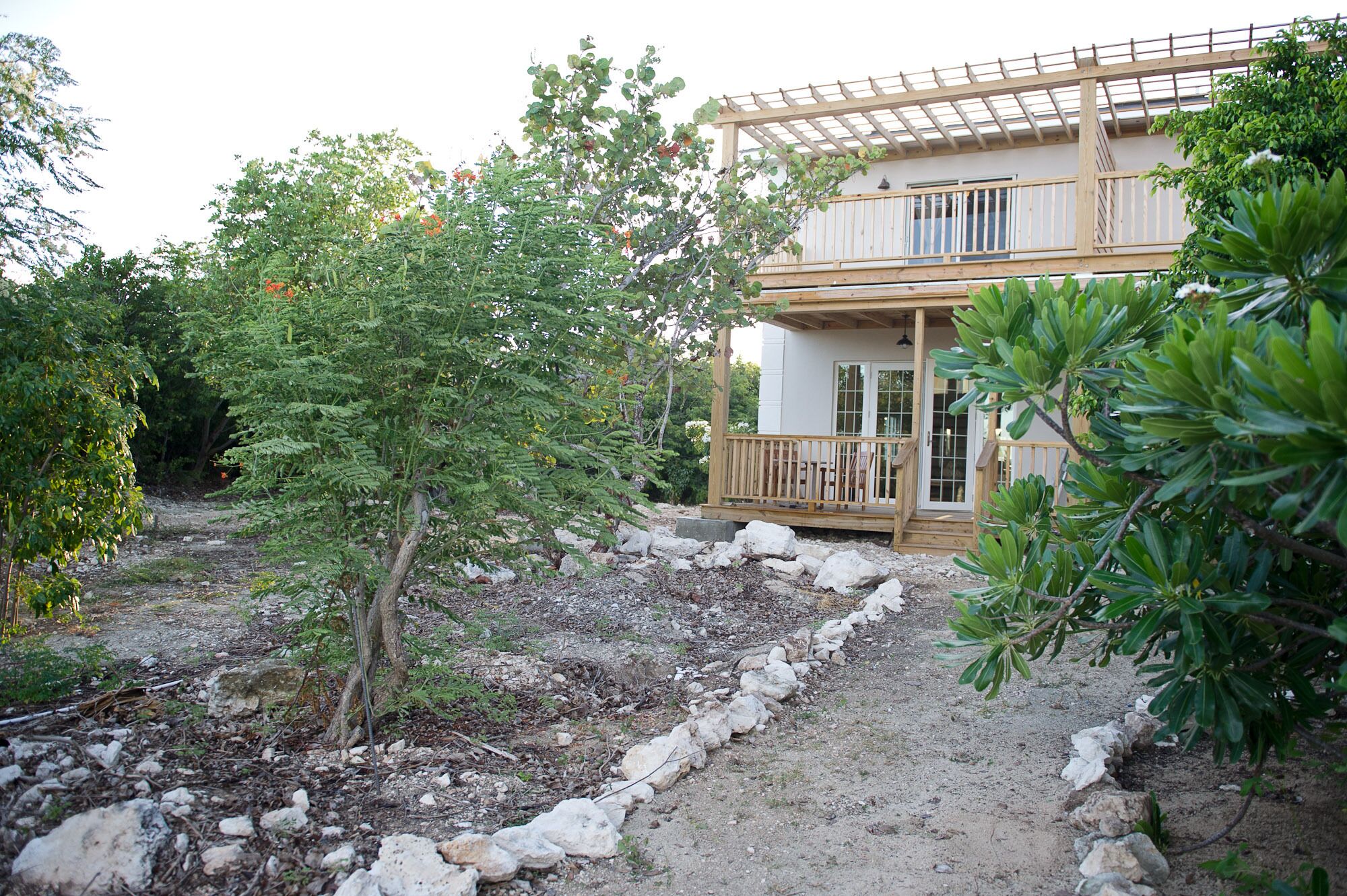 A two-story wooden house with a balcony, surrounded by a stone pathway and greenery.