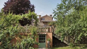 Une maison de campagne en pierre, avec une porte verte, un petit balcon et un jardin agrémenté de diverses plantes.
