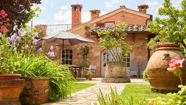 Une terrasse en pierre avec des plantes en pot, une table avec des chaises et un grand parasol.