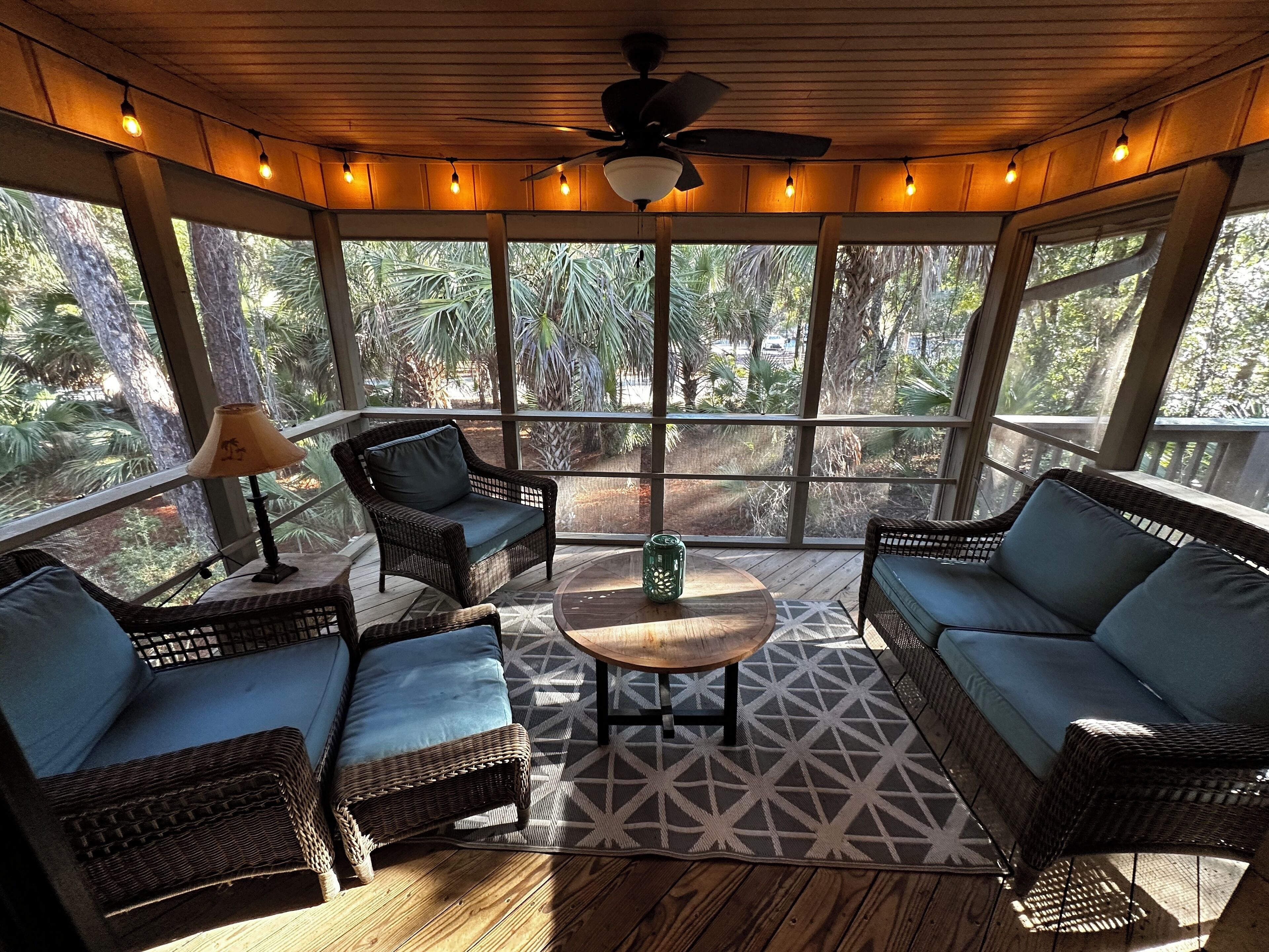 A covered outdoor seating area with wicker furniture, a ceiling fan, and large windows offering a view of the surrounding greenery.