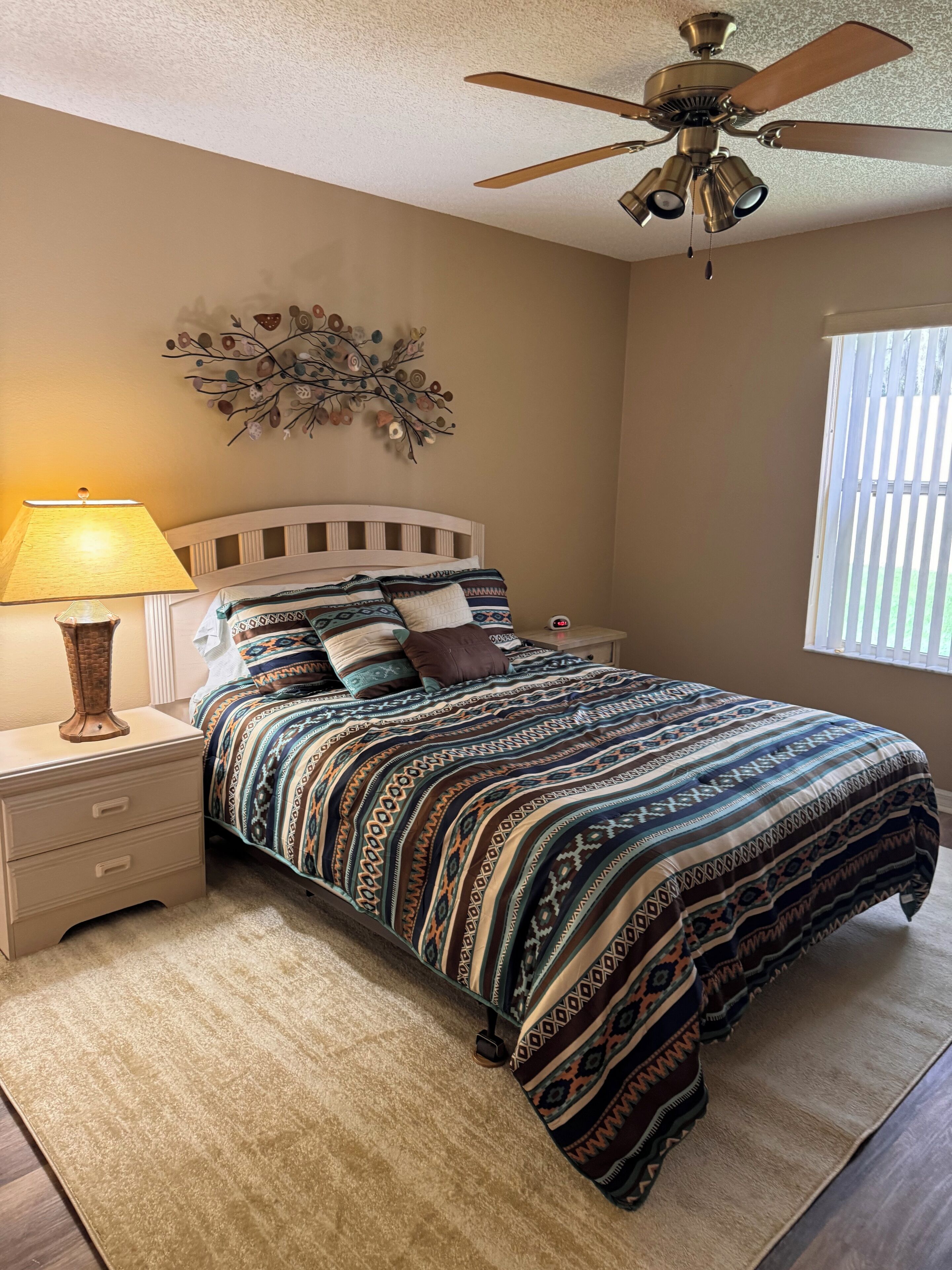 A bedroom with a bed, nightstand, lamp, ceiling fan, decorative wall art, and a window with blinds.