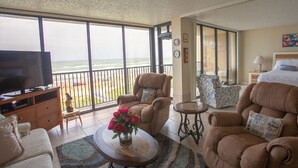 Ein geräumiges Wohnzimmer mit großem Fernseher, bequemen Sitzmöglichkeiten, einem Couchtisch mit Blumen und einem Balkon mit Meerblick.