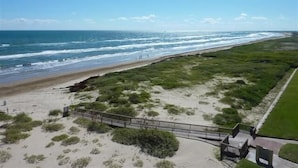 Blick auf die Küste mit einem Sandstrand, einer Grünfläche und einem Holzsteg, der zu einem Gebäude führt.