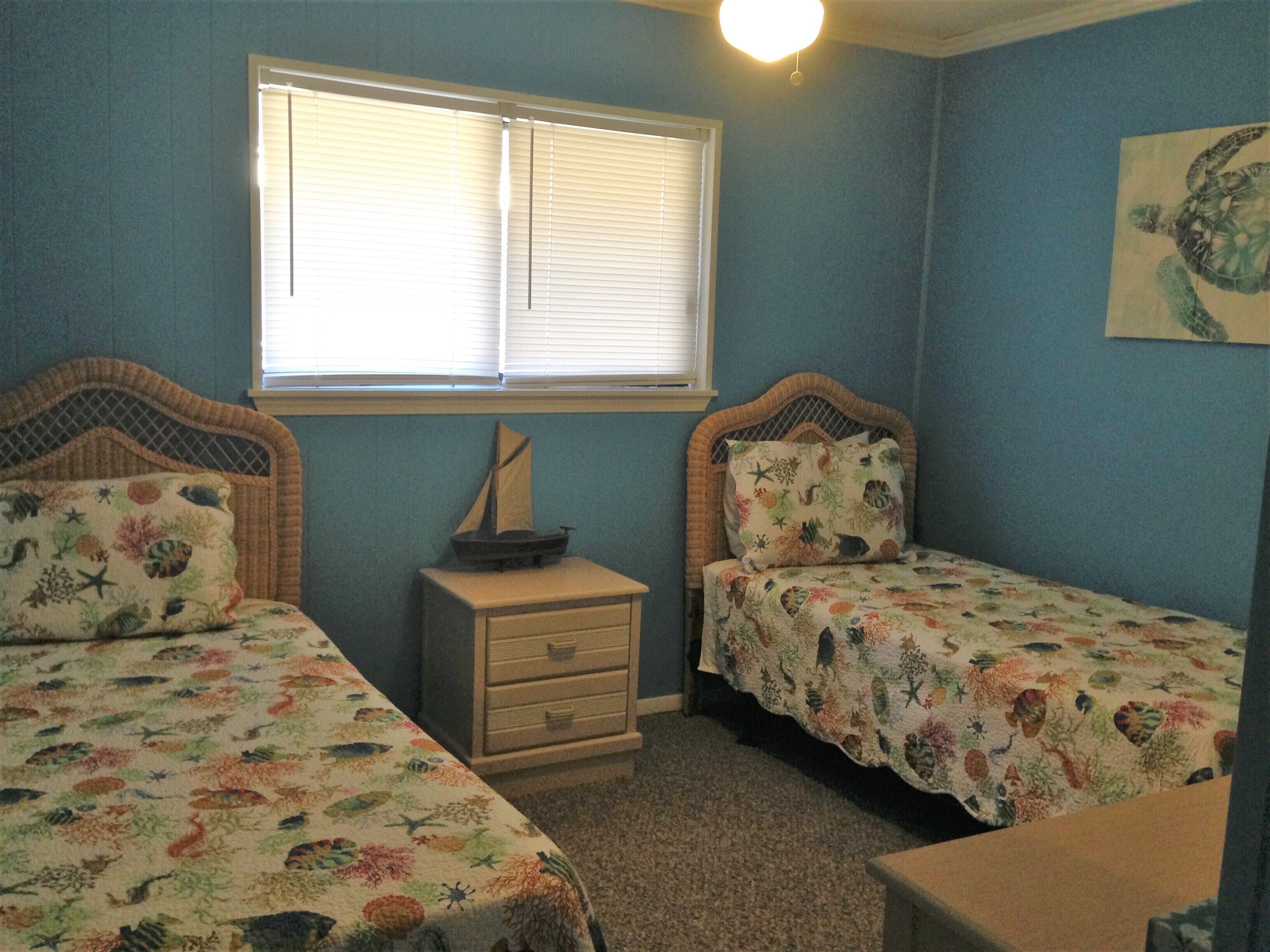 A bedroom with two single beds, a dresser, a window with blinds, and a small table.
