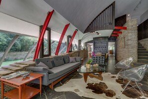 A modern living room with a sofa, glass coffee table, and clear chairs. A staircase with red accents leads to an upper level.