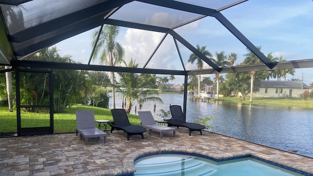 Área interna de piscina com vista para um lago, cercada por palmeiras e um alpendre coberto com espreguiçadeiras.