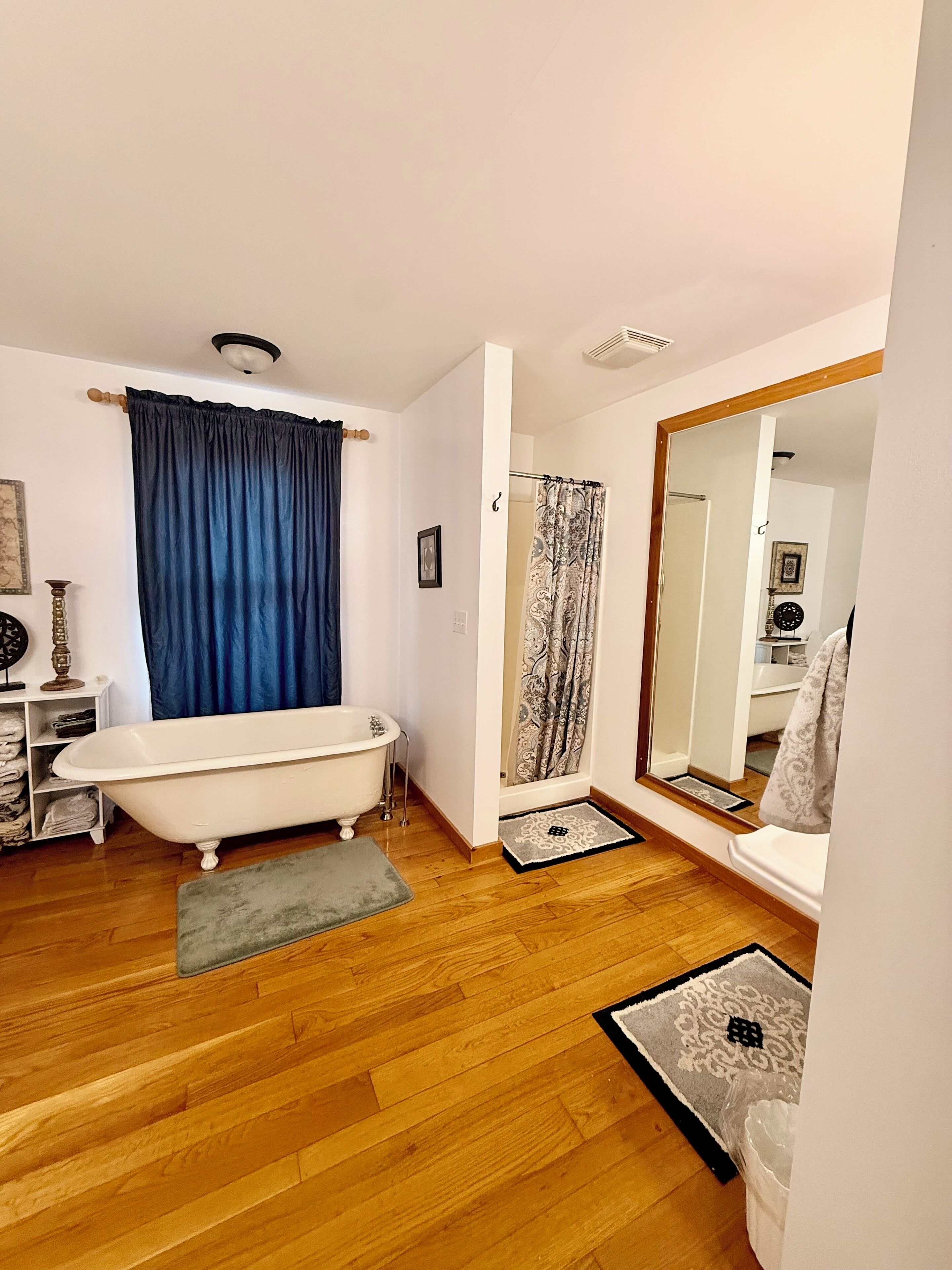 A bathroom with a white bathtub, blue curtains, a shower with patterned curtains, wooden flooring, and a large mirror.