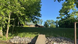 A wooden dock extending into a calm body of water, surrounded by lush greenery and a grassy hill with a house in the background. - Beautiful, Cedar Lakeside Cabin with Private Dock (Cadiz)