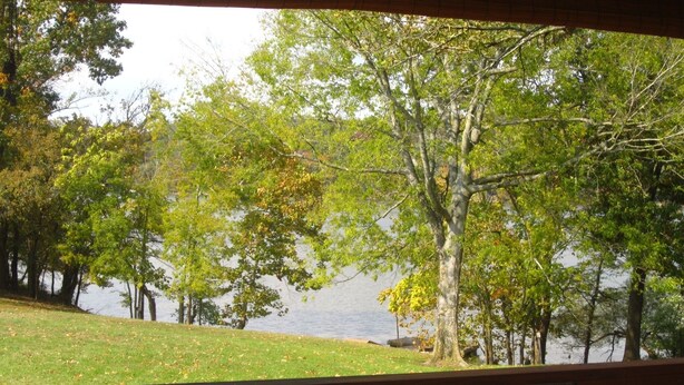 A view from a covered deck overlooking a lake with trees and grassy areas. - Beautiful, Cedar Lakeside Cabin with Private Dock (Cadiz)