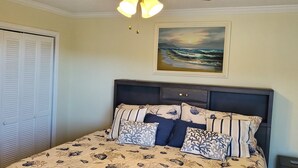 A bedroom with a bed, a nightstand, a painting of a beach scene, and a closet. - Beautiful 2 Bedroom, 2 Bath Ocean View Condo in Surfside Beach (Surfside Beach)