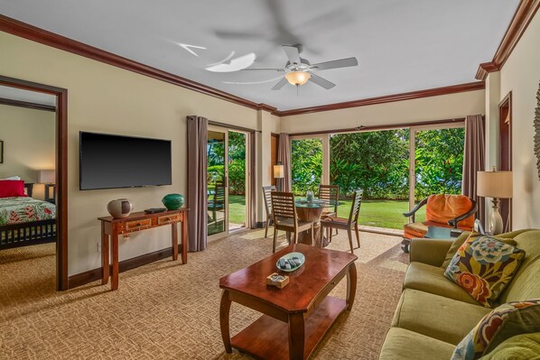 Amplia sala de estar con ventilador de techo, televisor de pantalla plana, comedor con mesa y sillas, y ventanas amplias que ofrecen vistas al exterior.