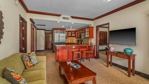 A living room with a sofa, coffee table, and a TV. There is a kitchen area with a refrigerator, stove, and bar stools. - Waipouli Beach Resort - Ground Floor #E103,  2 King Masters Suites (Kapaa)