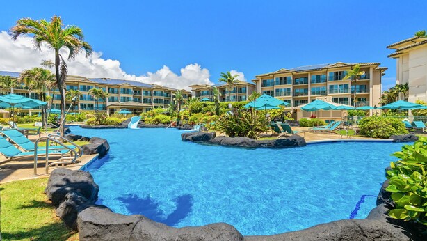 A swimming pool with lounge chairs and umbrellas, surrounded by a building with multiple balconies and a clear blue sky. - Waipouli Beach Resort - Ground Floor #E103,  2 King Masters Suites (Kapaa)