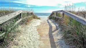 Une promenade en bois qui mène à la plage, avec du sable et de l’herbe de chaque côté.