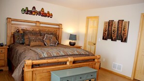 Ein Schlafzimmer mit einem hölzernen Bett, gemusterten Betttextilien, einem Nachttisch mit Lampe, einer kleinen Kommode und Wanddekoration.
