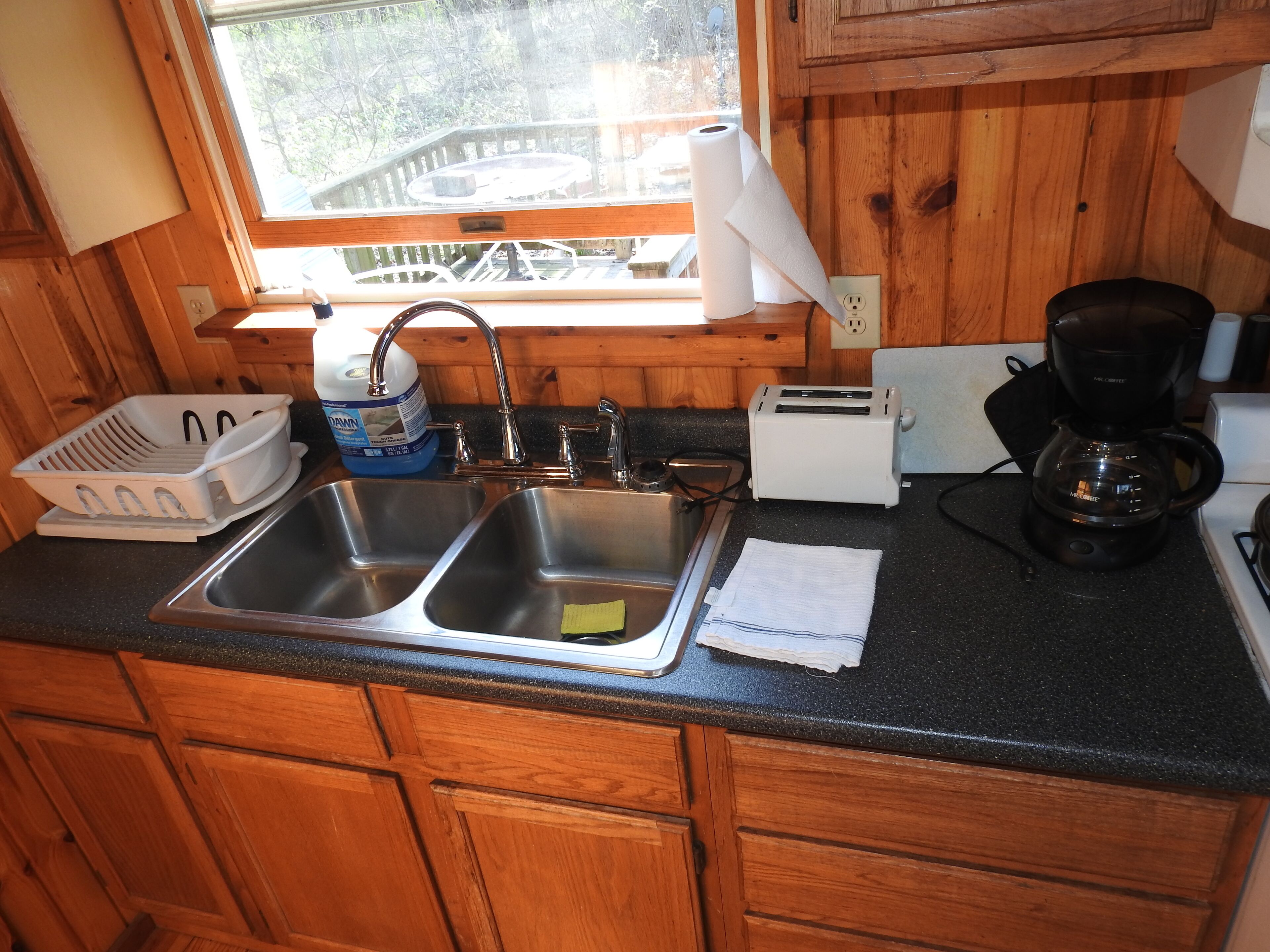 Une cuisine avec des meubles de rangement en bois, un évier à deux bacs, un grille-pain, une machine à café et une fenêtre donnant sur une terrasse.
