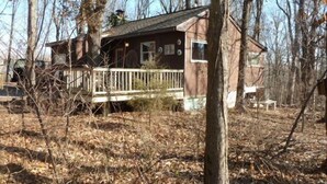 A rustic cabin with a wooden deck, surrounded by leafless trees and a dry ground.