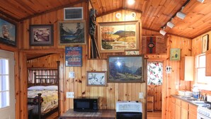 A cabin interior with wooden walls, a bunk bed, a kitchenette, and various framed pictures.