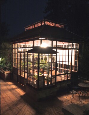 A glass-enclosed structure with a roof, surrounded by potted plants and outdoor furniture.