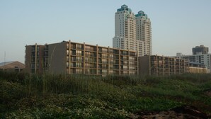 A multi-story building with a grid-like facade, surrounded by greenery and other buildings in the background.