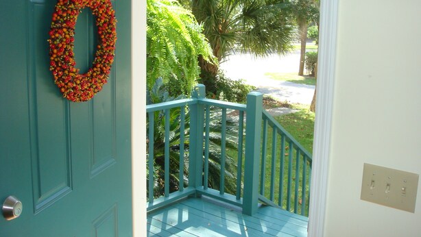 Une porte de couleur turquoise ornée d’une guirlande, une véranda bleue avec des balustrades et une vue sur une rue bordée de palmiers.