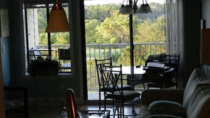 Un comedor con mesa y sillas, un sofá, y vista a un paisaje verde a través de amplias ventanas.