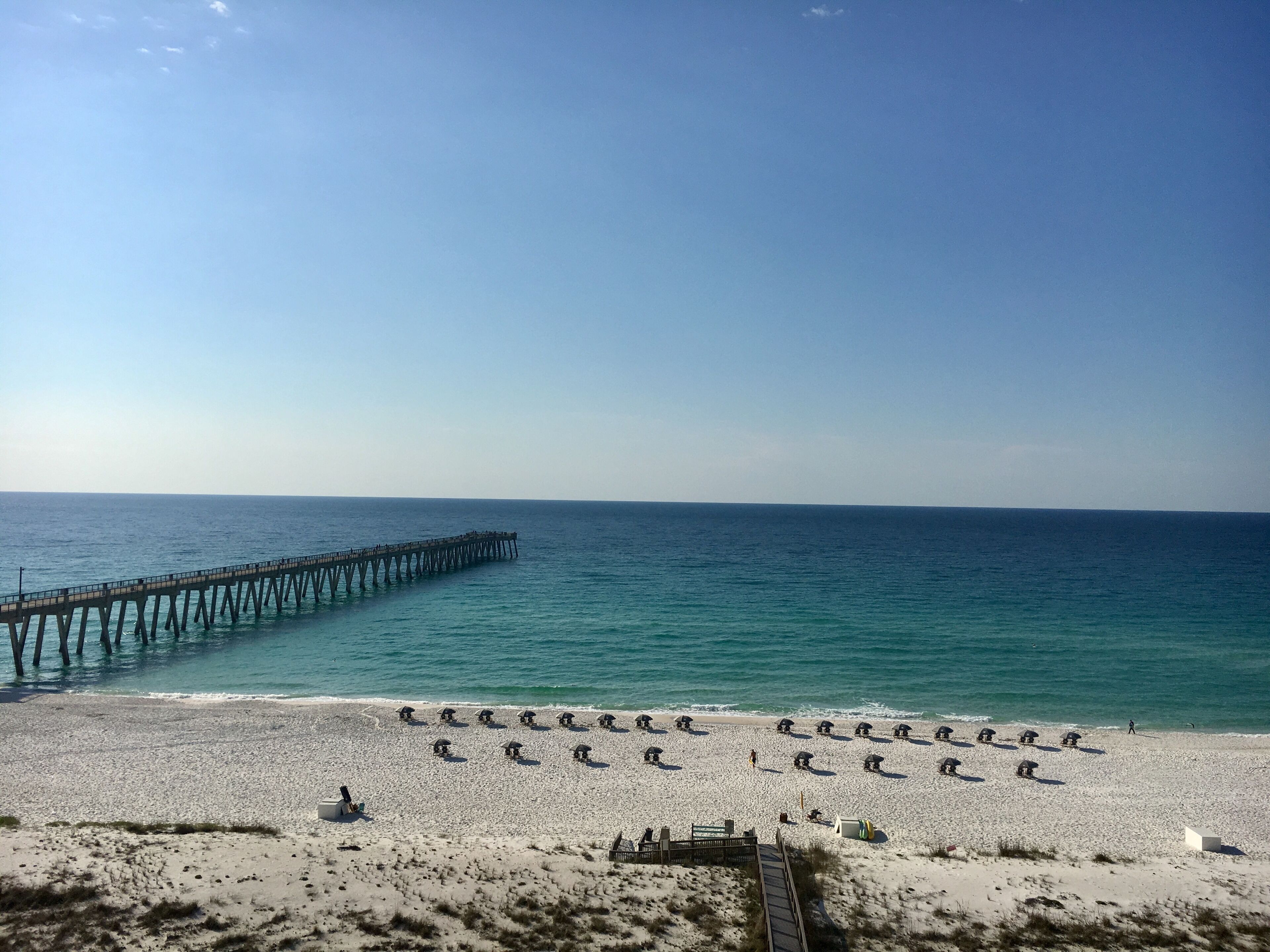 A beach with a long pier extending into the ocean, rows of beach umbrellas, and a sandy shoreline.