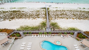 Vista aérea de un complejo turístico en la playa con un muelle largo, una zona de piscina y gente disfrutando de la playa.
