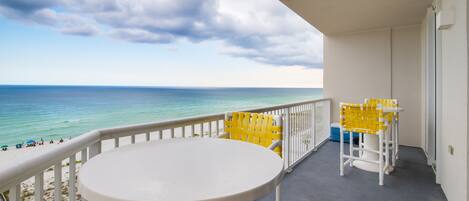 A balcony with a white table and yellow chairs, overlooking a beach with a clear view of the ocean.