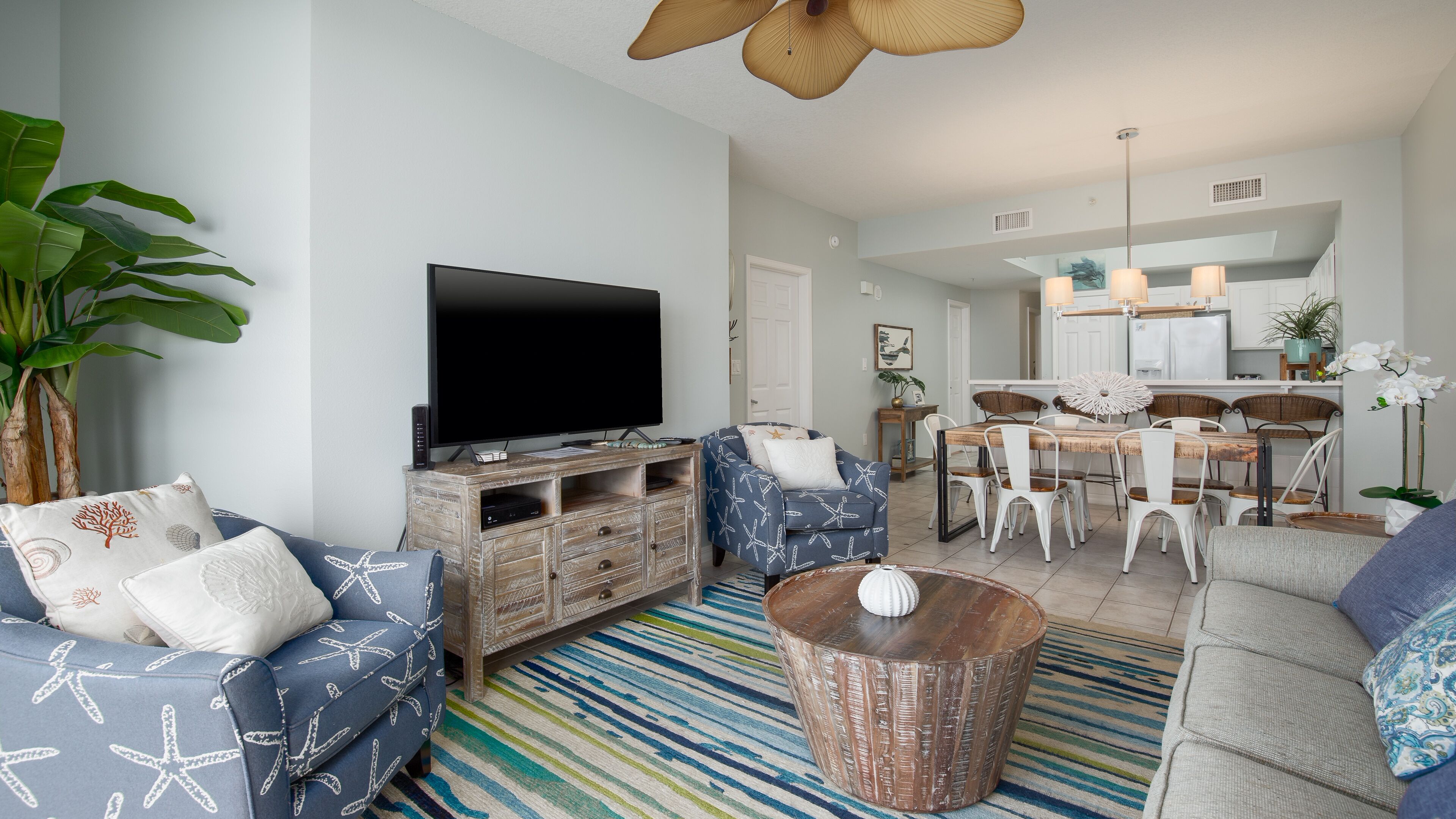 A modern living room with a flat-screen TV, blue patterned sofas, a coffee table, and a dining area with a wooden table and chairs.