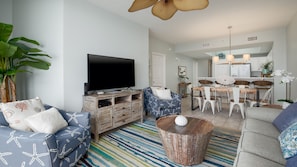 Una sala de estar moderna con televisor de pantalla plana, sofás azules con estampado, una mesa de centro y una zona de comedor con mesa y sillas de madera.