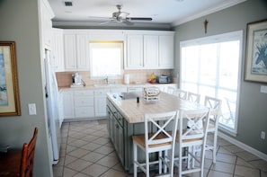 Une cuisine avec des meubles blancs, un sol carrelé, un ventilateur de plafond et un coin repas avec des chaises et une table blanches.