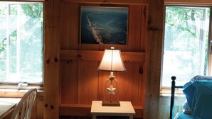 Een houten kamer met een bed, een bureau en een stoel. Aan de muur hangt een schilderij en op het bureau staat een lamp.