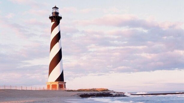 A lighthouse with a striped pattern, located on a sandy beach with the ocean in the background.