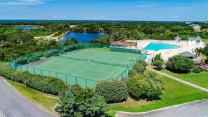A tennis court surrounded by lush greenery and a swimming pool with a deck area.