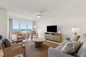 A spacious living room with a large TV, comfortable seating, a ceiling fan, and a view of the ocean.