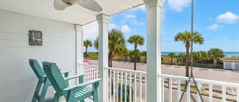 A covered porch with two green chairs, a ceiling fan, and a view of the beach and palm trees.