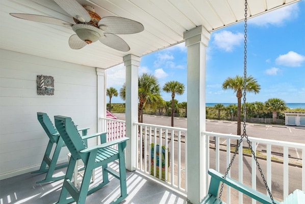 A covered porch with two green chairs, a ceiling fan, and a view of the beach and palm trees.