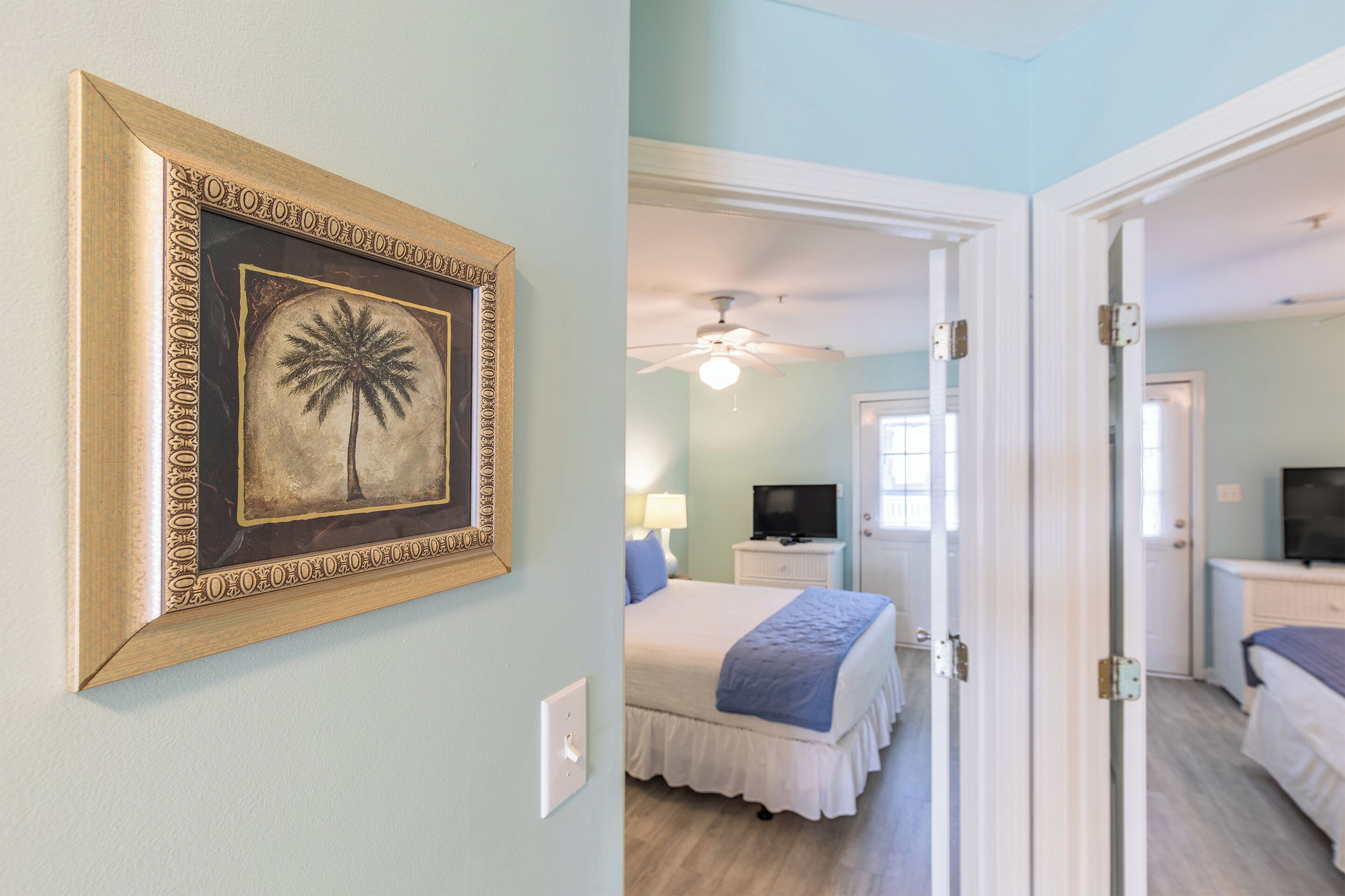 A room with a bed, a ceiling fan, a television, and a door leading to another room.