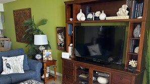 A living room with a large flat-screen TV, a blue sofa, a coffee table, and a wicker ottoman.