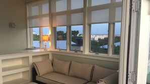 A room with a beige sectional sofa and a matching ottoman, large windows with blinds, and a lamp on the windowsill. - 5 BR, 5 BA in Seaside, quick walk to the private beach access or main pool  (Santa Rosa Beach)