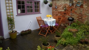 Pieni patio, jossa on pöytä ja tuoleja, tiiliseinä ja roikkuvia kukkaruukkuja.