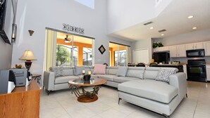 A spacious living room with a large L-shaped gray sofa, a round wooden coffee table, and a modern kitchen in the background. - TOP REVIEWED HOME! Only 3 MILES to Disney, South Pool in Emerald Island Resort (Kissimmee)