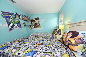 A bedroom with a large bed covered in a Buzz Lightyear-themed blanket, a wall with various animated character posters, and a nightstand with a lamp.