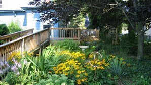 A garden with various plants and flowers, a wooden deck, and a stone fountain.