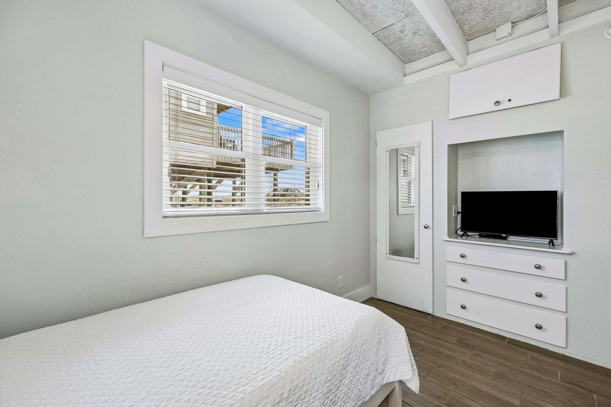 Un dormitorio con cama, televisión, ventana con persianas y espejo.