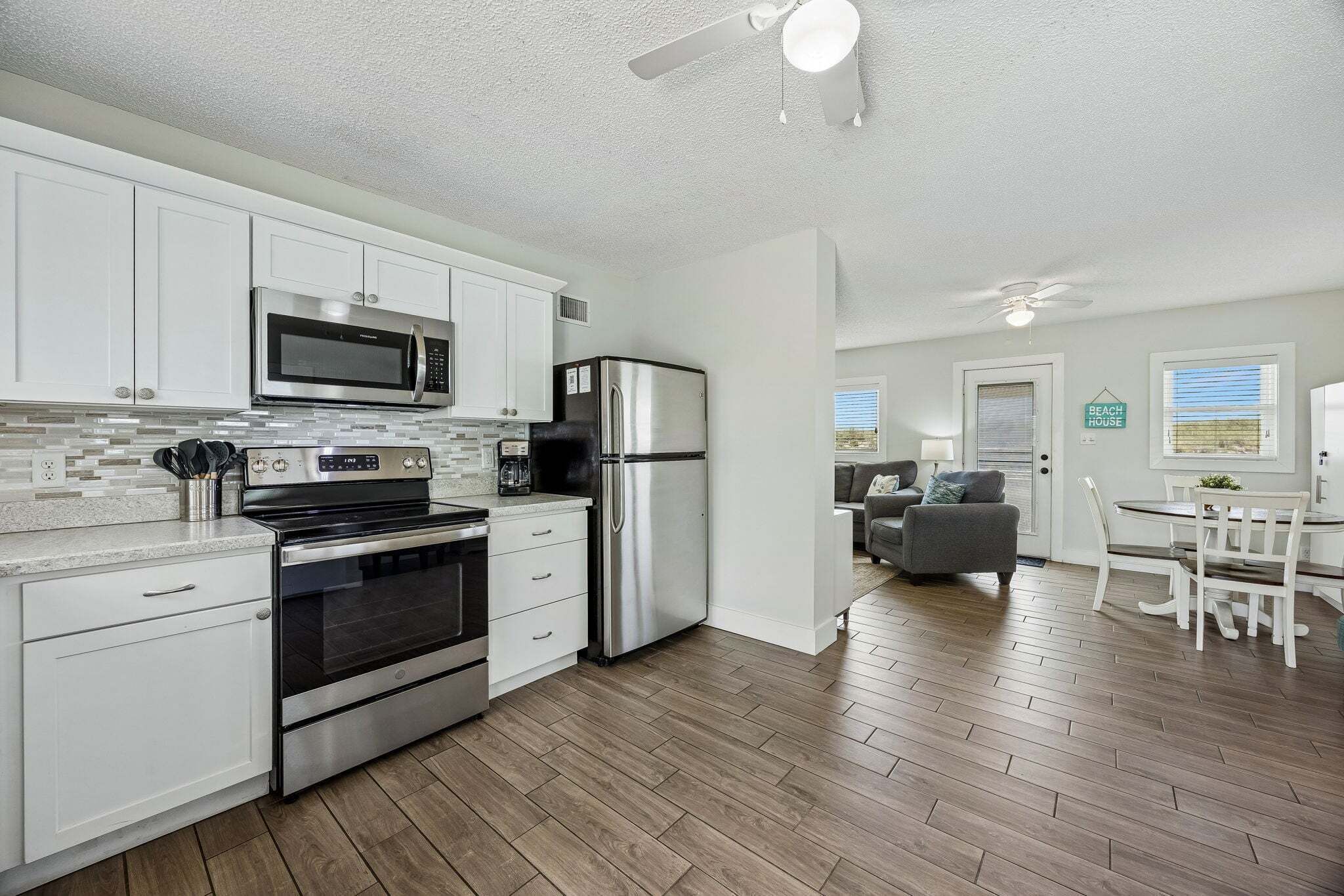 A modern kitchen with stainless steel appliances, a dining area with a white table and chairs, and a living room with a gray sofa.