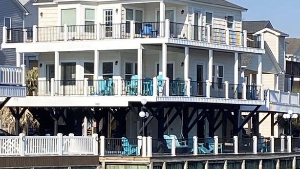 Un bâtiment situé au bord de l’eau, doté de nombreux balcons, d’un quai et d’un espace extérieur aménagé pour s’asseoir.