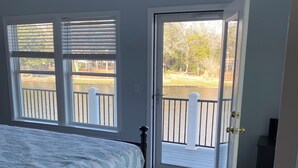Une chambre avec un grand lit, deux fenêtres équipées de stores, une porte donnant sur un balcon et une vue sur une étendue d’eau et des arbres.